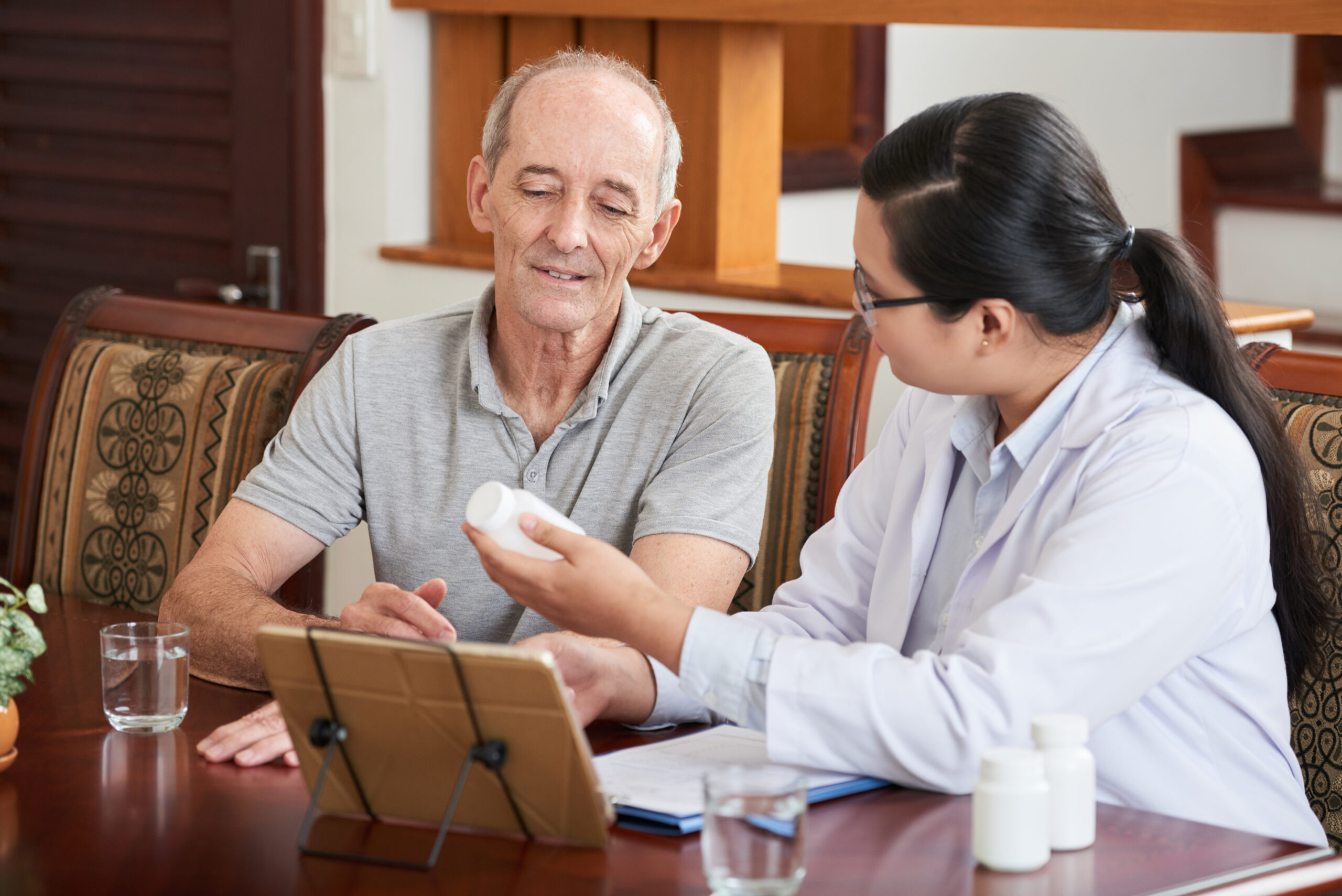 Woman at table with tablet and papers and showing pills in jar to senior patient sitting near