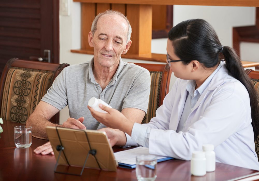 Woman at table with tablet and papers and showing pills in jar to senior patient sitting near