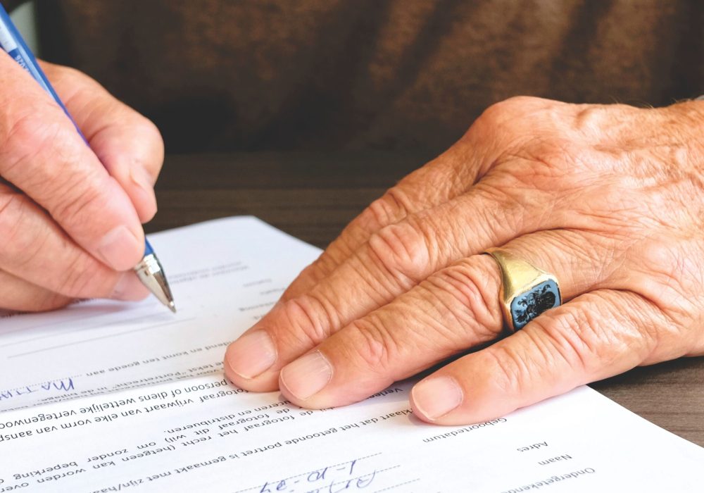 Close-up of a senior adult signing a legal document with a focus on hand and gold ring.